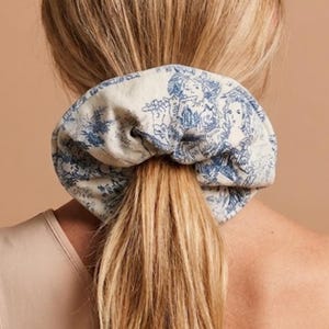 Op de afbeelding: Een crèmekleurige scrunchie met een blauwe toile de Jouy-print, met figuren die muziekinstrumenten bespelen. De scrunchie is om een blonde paardenstaart gewikkeld. De achtergrond is effen en neutraal.