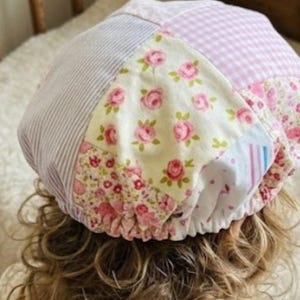May include: A patchwork baby bonnet with various floral, striped, and gingham patterns in pink, white, and lavender. The bonnet features a ruffled edge and is designed to fit a baby's head.