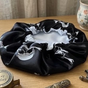 Pode incluir: Touca de cabelo de cetim preto com um padrão de caveira e ossos cruzados brancos. O interior é branco. A touca foi projetada para proteger o cabelo durante o sono. O design de caveira e ossos cruzados dá-lhe um tema gótico ou pirata.