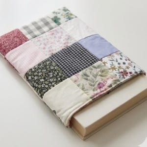 Op de afbeelding: Een patchwork boekomslag met verschillende stofpatronen, waaronder bloemen, ruitjes en effen kleuren. De omslag is ontworpen voor een boek, met de rug en pagina's zichtbaar. De omslag is gemaakt van zachte stof.