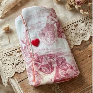 May include: A fabric case with a red and white toile design, folded and secured with a small red heart. The case, possibly for small items, has a vintage style and feel.
