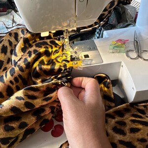 May include: A close-up of a sewing machine with a leopard print fabric being sewn. The fabric is being held in place with clips and the machine is running.