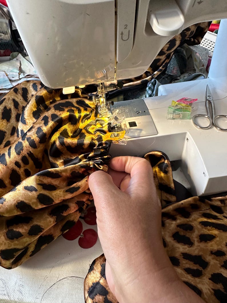 May include: A close-up of a sewing machine with a leopard print fabric being sewn. The fabric is being held in place with clips and the machine is running.