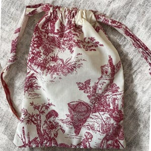 Puede incluir: Una bolsa con cordón con fondo crema y un estampado toile de Jouy burdeos. El estampado representa una escena pastoril con figuras, árboles y edificios. La bolsa tiene un cierre de cordón a juego.