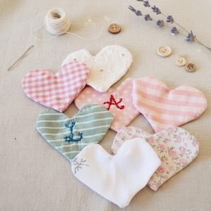 Puede incluir: Una colección de adornos de tela en forma de corazón en varios patrones y colores, incluyendo cuadros rosas, encaje blanco e estampados florales. Algunos corazones presentan detalles bordados como una ancla o la letra "A". También hay botones, una aguja y un carrete de hilo.