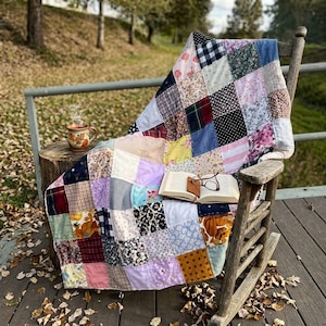 Peut inclure: Une courtepointe en patchwork composée de divers carrés de tissu aux couleurs et motifs variés, incluant des imprimés floraux, à carreaux et vichy. La courtepointe est drapée sur un fauteuil à bascule en bois, avec un livre ouvert et une tasse.