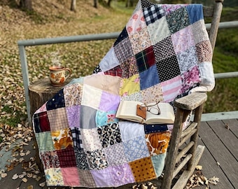 May include: A patchwork quilt made of various fabric squares in diverse colors and patterns, including floral, plaid, and gingham prints. The quilt is draped over a wooden rocking chair, with an open book and a ceramic mug resting on the seat.