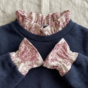 May include: A dark navy blue long-sleeved sweater featuring a cream-colored ruffled collar and matching ruffled sleeve cuffs. The ruffles are decorated with a detailed maroon-colored toile pattern depicting outdoor landscape scenes.