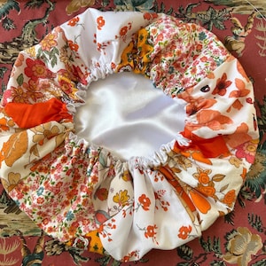 Könnte beinhalten: Eine Patchwork-Duschhaube mit weißem Satin-Innenfutter. Die Außenseite zeigt florale Muster in Orange, Gelb und Rosa auf weißem Hintergrund. Die Haube hat einen elastischen Rand.