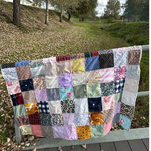 Può includere: Una trapunta patchwork realizzata con diversi tessuti di colori e motivi diversi. La trapunta è composta da quadrati di tessuto cuciti insieme per creare un design più grande. La trapunta è appesa a una ringhiera di legno.