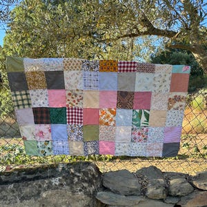 以下が含まれることがあります： 様々な色と柄の生地の正方形を組み合わせたパッチワークキルト。花柄、チェック柄、無地など。キルトはフェンスに掛けられており、背景には木と空の自然な風景が広がっています。