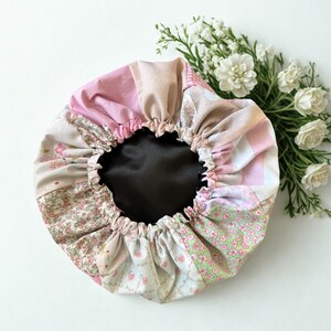 Puede incluir: Un gorro forrado de satén con patchwork en tonos rosa, crema y verde. El gorro presenta una variedad de telas estampadas florales, con un borde elástico fruncido y un interior oscuro. Flores blancas en el fondo.