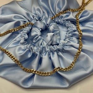 Pode incluir: Uma bolsa de cetim azul claro com cordão e corrente dourada. A bolsa tem uma parte superior franzida e uma textura brilhante e lisa. A corrente dourada é grossa e adiciona um toque de elegância ao acessório.