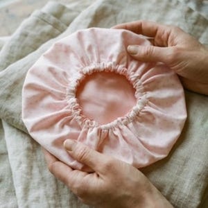 May include: A pink shower cap with a white polka dot pattern. The cap has an elastic band around the edge and is held by hands. The interior is a solid pink color. The cap is made of a soft fabric.