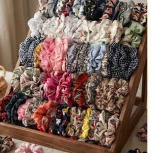 Puede incluir: Un expositor de madera lleno de coleteros coloridos. Los coleteros presentan diferentes patrones y colores, incluyendo florales, lunares y colores lisos. Los accesorios para el cabello están dispuestos en filas ordenadas, mostrando una amplia selección.