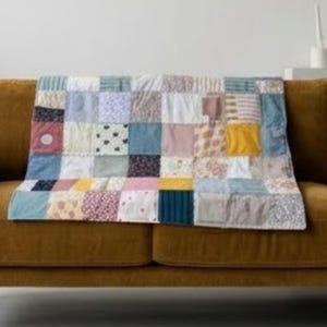 May include: A rectangular patchwork quilt featuring a grid of fabric squares in various colours and patterns, including shades of blue, yellow, pink, white, and grey, with floral, polka dot, striped, and solid designs arranged in a multi-coloured layout.