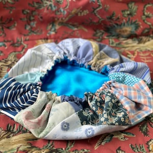 以下が含まれることがあります： パッチワークのデザインが施された、丸い青い布製のクッション。クッションは、さまざまな青、白、ピンクの色合いの布の異なる部分で作られています。クッションは、柔らかく青い素材で満たされています。
