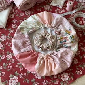May include: A handmade pink and floral fabric hair bonnet and matching scrunchie. The bonnet is made of various pink and floral patterned fabrics. The scrunchie is made of a matching floral fabric. The items are displayed on a red floral fabric.