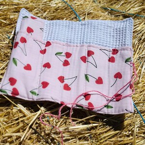 Könnte beinhalten: Ein rosa Stoffartikel mit Kirschmuster, mit roten Kirschen und grünen Blättern auf hellrosa Hintergrund. Der Artikel ist mit einem weißen Waffelstoff kombiniert. Rosa Faden ist sichtbar, was auf einen handgefertigten Artikel hindeutet.