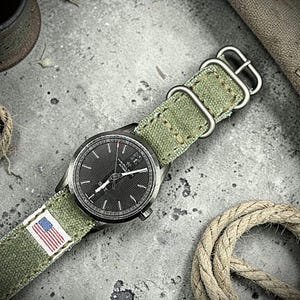 May include: A Hamilton automatic wristwatch with a round black dial featuring silver indices and hands. The watch is attached to a textured olive green canvas strap with a small American flag patch and silver-toned metal hardware buckles.