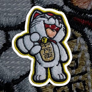 Könnte beinhalten: Gestickter Aufnäher mit einer weißen Maneki-neko-Katze mit roten Akzenten, die eine goldene Münze hält. Die Katze hat einen fröhlichen Ausdruck, umrandet in Schwarz und Gelb. Die Münze hat japanischen Text.