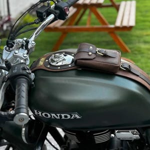 Op de afbeelding: Close-up van een donkergroene Honda motorfiets met een bruine leren tankhoes en een kleine bijpassende tas. Het stuur, de meters en het woord "HONDA" op de tank zijn zichtbaar.