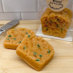 May include: Orange-colored zucchini bread soy wax melts with green flecks, displayed on a wooden cutting board. The Waxy Bliss Co. label is visible on the packaged bread. A silver butter knife is also present.