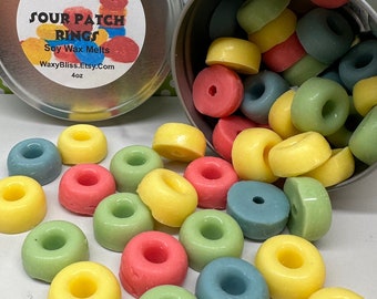 May include: A silver tin with a label reading "Sour Patch Rings Soy Wax Melts" next to a spilled tin of colorful, ring-shaped wax melts. The melts are in shades of red, yellow, green, and blue.