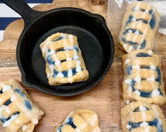 May include: Several miniature blueberry pies with lattice crusts are arranged on a wooden serving board. One pie sits in a small black cast iron skillet. The pies have a light brown crust, blue filling, and white icing.