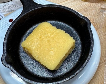 May include: A close-up of cornbread-shaped soy wax melts. One melt is in a miniature cast iron skillet, and three are packaged in clear plastic. The packaging includes the text "Cornbread Soy Wax Melts" and a website address.
