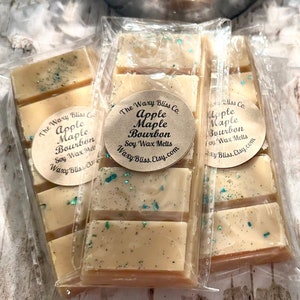 May include: Several soy wax melt bars in clear packaging. Each bar is light beige with flecks of blue and green. The bars are labeled "Apple Maple Bourbon" from "The Waxy Bliss Co."
