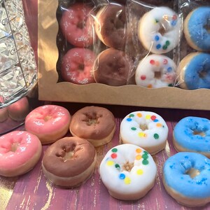 May include: A collection of colorful donuts, some in a clear box, others displayed separately. The donuts feature pink, brown, white, and blue frosting with sprinkles. The box is brown with a clear window, and the donuts are arranged on a pink surface.