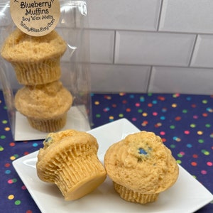 May include: Soy wax melts in the shape of blueberry muffins. The light brown muffins have blue accents and are displayed on a white square plate. The packaging includes a clear bag with three muffins and a label that reads "Blueberry Muffins Soy Wax Melts".