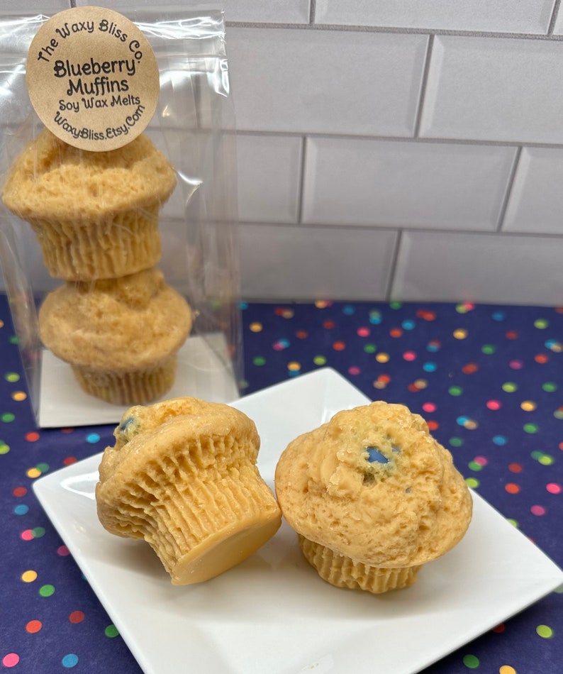 May include: Soy wax melts in the shape of blueberry muffins. The light brown muffins have blue accents and are displayed on a white square plate. The packaging includes a clear bag with three muffins and a label that reads "Blueberry Muffins Soy Wax Melts".