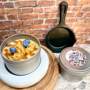 May include: A soy wax candle in a silver tin, topped with waffle-shaped wax and blue wax blueberries, drizzled with caramel-colored wax. A small cast iron skillet and a second candle in a silver tin are also present.