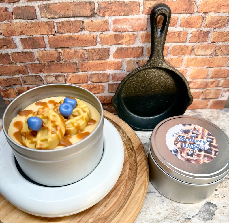 May include: A soy wax candle in a silver tin, topped with waffle-shaped wax and blue wax blueberries, drizzled with caramel-colored wax. A small cast iron skillet and a second candle in a silver tin are also present.