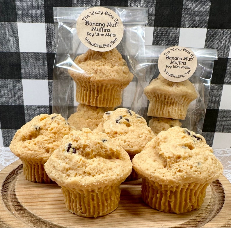 May include: Several banana nut muffins, some in clear bags, displayed on a wooden board. The muffins are a light brown colour with dark specks. The bags have labels that read "Banana Nut Muffins Soy Wax Melts".