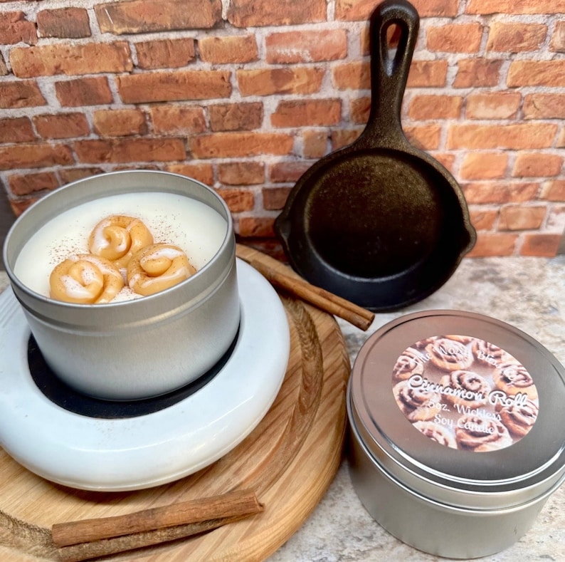 May include: A silver tin candle with a white wax base and three cinnamon roll-shaped wax decorations. The candle sits on a white warmer, with a second candle in a silver tin featuring a cinnamon roll label. A small cast iron skillet and cinnamon sticks are also present.