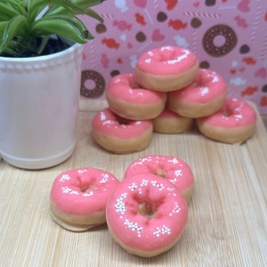 May include: A collection of miniature donuts with pink frosting and white sprinkles. The donuts are arranged on a wooden cutting board, with a white ceramic pot holding a green plant in the background. The donuts have a light brown base.