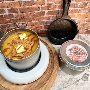 May include: A candle in a silver tin, designed to look like French toast, with a small cast iron skillet and a second candle. The French toast candle has a yellow wax base with a brown drizzle and two square butter-shaped wax pieces.