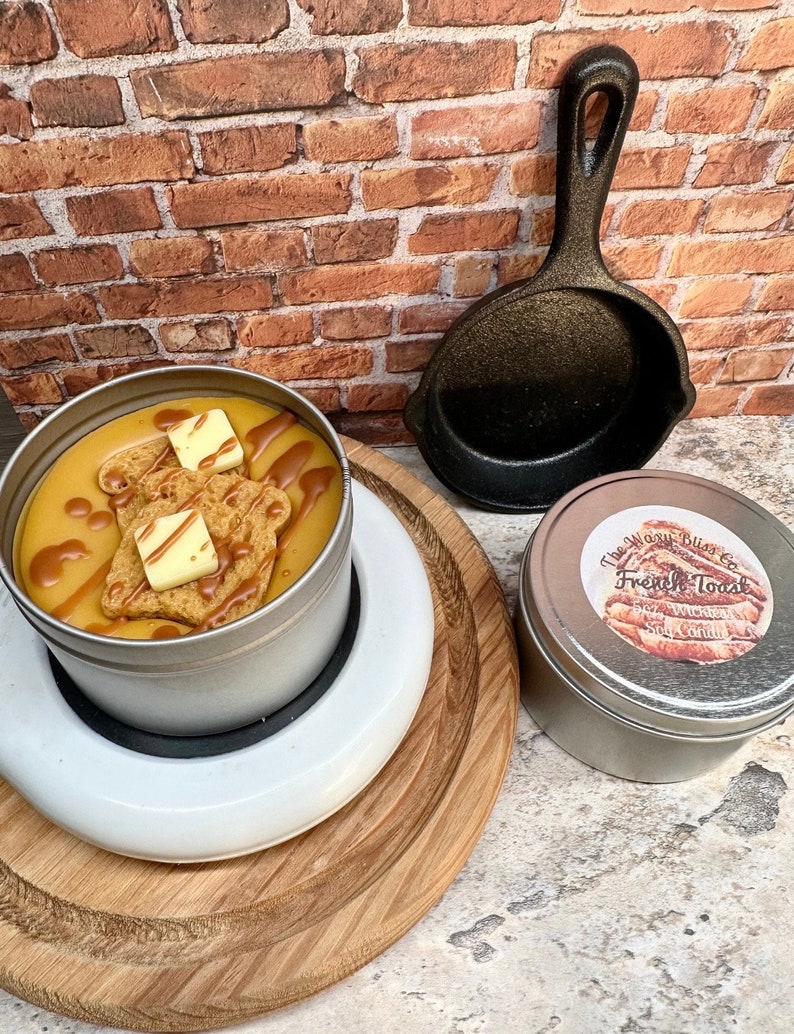 May include: A candle in a silver tin, designed to look like French toast, with a small cast iron skillet and a second candle. The French toast candle has a yellow wax base with a brown drizzle and two square butter-shaped wax pieces.