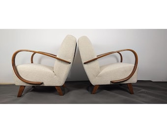 May include: Two mid-century modern armchairs. Each chair has a curved wooden armrest and legs, with a cream-colored, textured fabric seat and back. The chairs are positioned facing each other.
