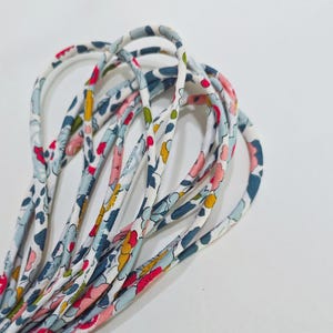 May include: A coiled length of fabric cord featuring a white base with a dense floral pattern in shades of pink, yellow, light blue, and dark blue. The cord is rounded and flexible, displaying a continuous repeating print of small blossoms.