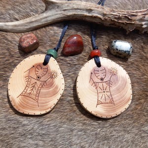 May include: Two wooden pendants with intricate designs, hanging from black cords. The pendants are oval-shaped and feature a stylized human figure with geometric patterns. The pendants are displayed on a brown fur background.