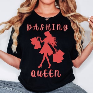 Puede incluir: Camiseta negra con una silueta roja de una mujer que lleva bolsas de la compra. El texto "Dashing Queen" está impreso en rojo en la camiseta.