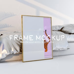 May include: A gold frame mockup with a white background and a pink border. The frame is leaning against a white bed with a silver satin duvet and pillows. The text "FRAME MOCKUP" is written in white on the frame, with the tagline "Elevate your prints & artworks" below it.