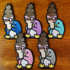 May include: Five embroidered iron-on patches featuring penguins holding ice cream cones. The penguins are in different colors: blue, purple, pink, gray, and teal. The ice cream cones are all brown with pink dripping ice cream.