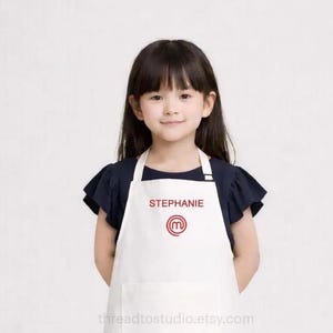 May include: A white apron featuring the name STEPHANIE in red text above a red circular logo with a lowercase m inside. The apron has a front pocket and is worn over a dark navy short-sleeved shirt and light beige trousers with white trainers.