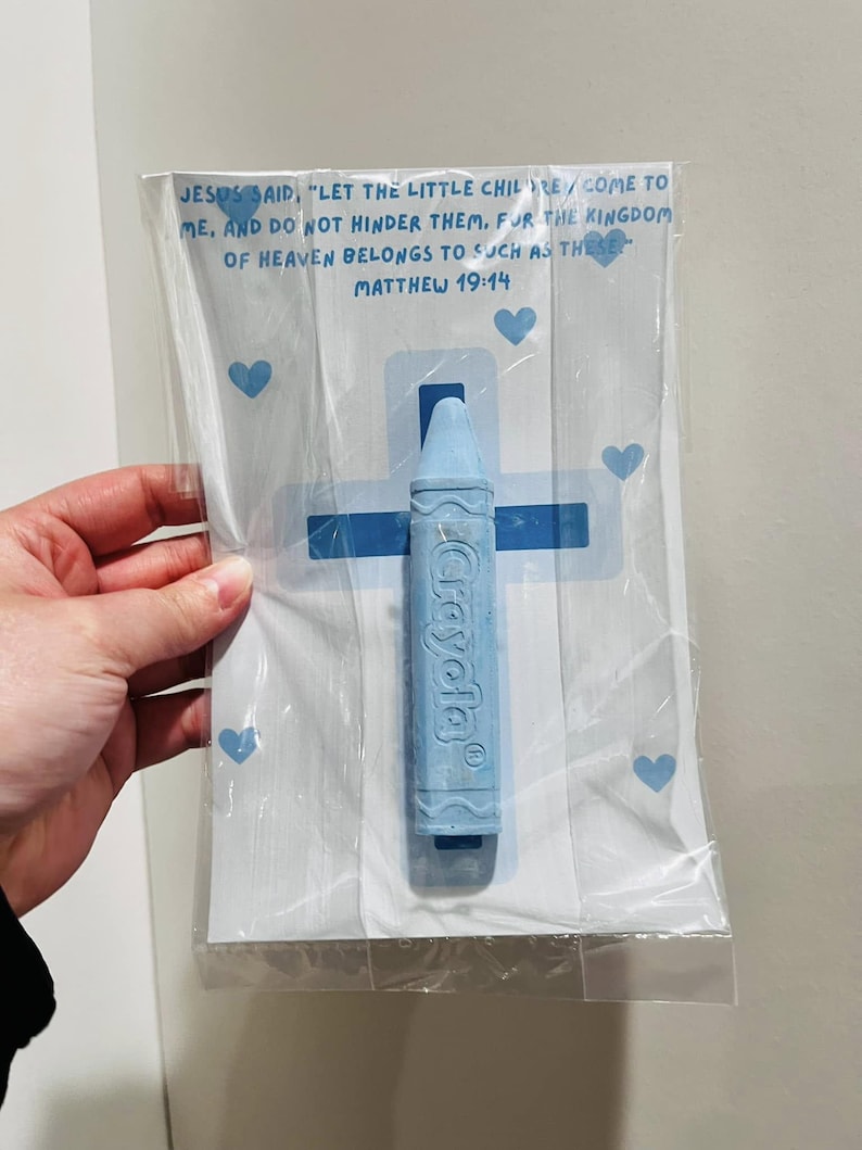 May include: A blue crayon with the word "Crayola" printed on it, packaged in a clear plastic bag with a white card. The card has a blue cross and hearts printed on it. The card also has a bible verse printed on it: "Jesus said, 'Let the little children come to me, and do not hinder them. For the kingdom of heaven belongs to such as these.' Matthew 19:14"