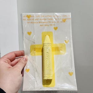 May include: A yellow crayon with the word "Crayola" printed on it, packaged with a white card featuring a yellow cross and hearts. The card includes a Bible verse from Matthew 19:14, "Let the little children come to me, and do not hinder them, for the kingdom of heaven belongs to such as these."
